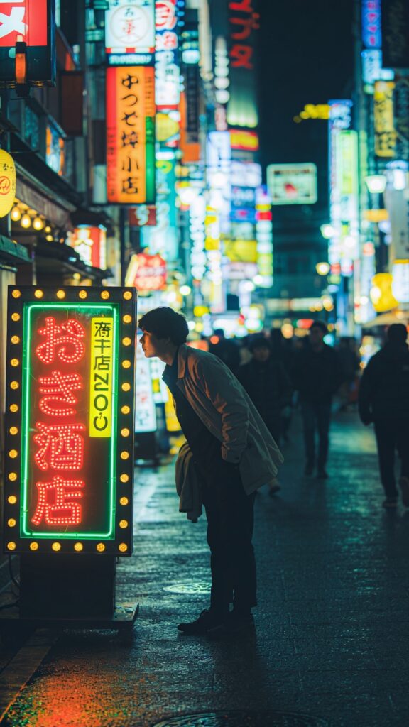 Create a high-resolution, cinematic street photo set at night in a bustling Japanese neon-lit district. The main subject should be a person standing slightly off to the side, leaning in as if whispering a secret to the glow of neon signs. The subject is rotated about 45 degrees to create a dynamic silhouette. Keep hands loose in pockets or draped casually over a light jacket. Use a low ISO to ensure the lights remain crisp and vibrant, with shallow depth of field that gently blurs background signage while retaining readable characters and color. The scene should feature vivid, multicolored signage in kanji and romaji, reflections on wet pavement, a shallow crowd in the background, and a cinematic contrast between the subject and the luminous signs. No text on the image. The composition should convey a sense of location and mood, with the neon glow casting cool and warm tones on the subject’s face and clothing, capturing a spontaneous, stylish travel snapshot in Japan at night.
