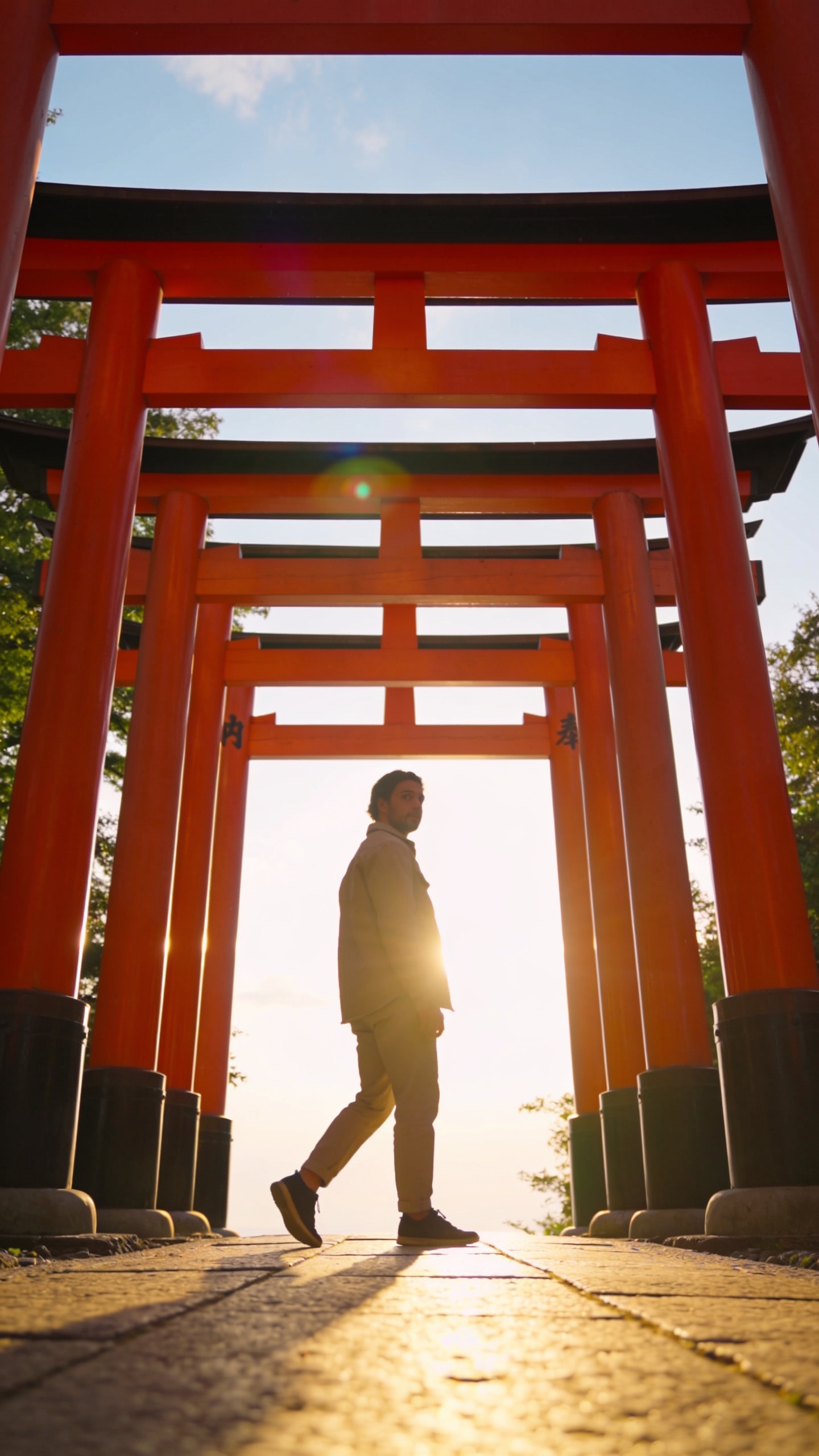A high-resolution, cinematic travel portrait of a solo traveler walking through a traditional torii gate at a Shinto shrine in Japan, captured in early golden hour light. The main subject stands with one foot stepping forward, then turns their upper body 90 degrees to the gate to create a crisp profile as they pause to look over their shoulder. The torii gate forms a strong, symmetrical frame around the subject, creating the effect of stepping into a painting. The scene is shot from a slight low angle to emphasize the gate’s vertical lines and create depth, with the horizon kept level to avoid distortion. The gate’s vermillion color contrasts with a soft blue sky and lush greenery in the background, while subtle lens flare adds warmth. The subject wears contemporary, clean attire (neutral tones) to keep the focus on the gate’s architecture, with natural, relaxed expression and a calm, iconic pose that instantly signals Japan. No text or logos in the image.