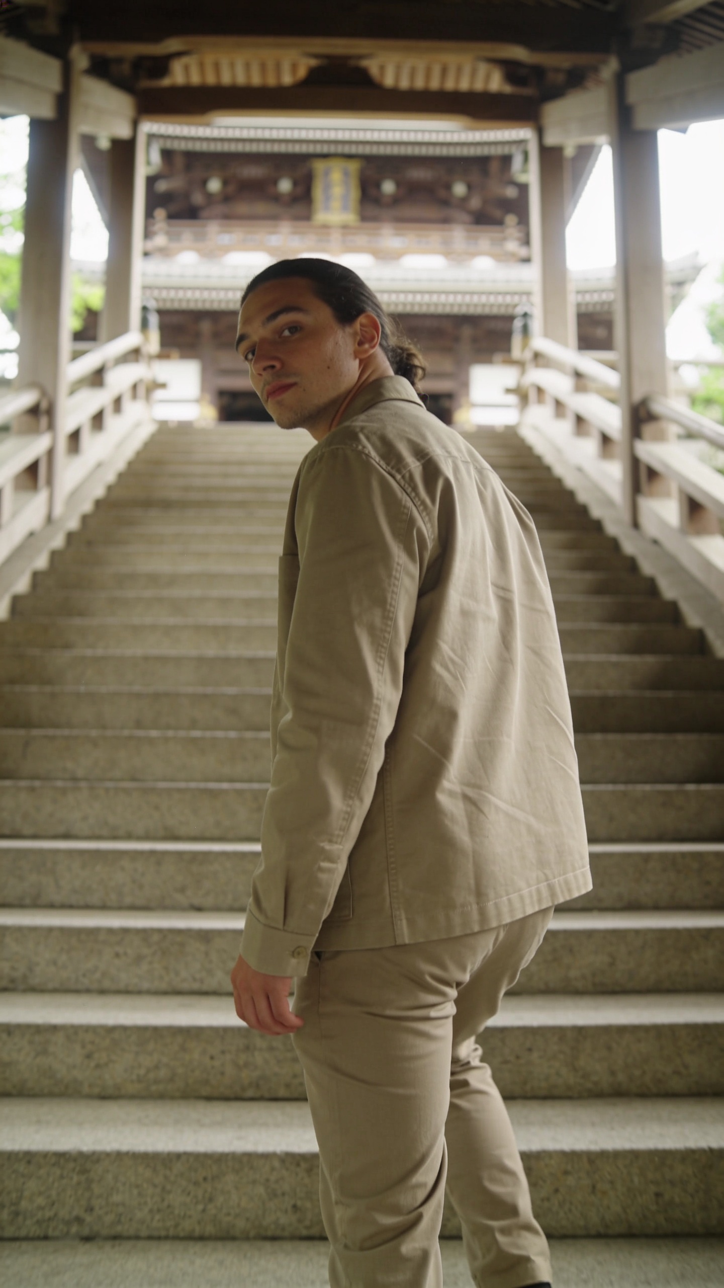 A cinematic, high-resolution portrait of a solo traveler posing on a grand stone staircase inside a serene temple setting in Japan. The subject is turned back over their shoulder, mid-climb, with a confident, cinematic gaze toward the camera. Soft, warm natural light from the side highlights the subject’s relaxed shoulders and natural chin tilt, creating gentle shadows that define facial features. The staircase lines guide the viewer’s eye up to the subject, while a shallow depth of field keeps the ornate temple architecture and distant, slightly blurred background subtly visible. The person wears a stylish, understated travel outfit—neutral-toned fabric with clean lines, a lightweight jacket, and comfortable pants—emphasizing a premium, timeless aesthetic. The scene exudes calm majesty, with textured stone steps, subtle greenery in the background, and a quiet, contemplative mood, as if capturing a moment of Zen and confidence. No text on the image.